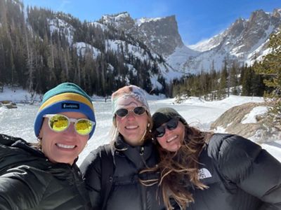 Three women stand in front of snow-blanketed mountains and valley.