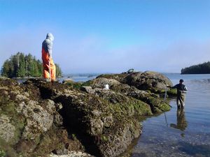 A man in waders stands knee-deep in ocean water while another person wearing waders stands atop a rock jetty.