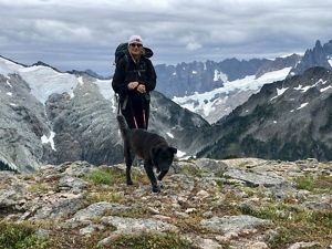 Martha Kongsgaard stands with a black dog on a leash atop a mountain with more of a mountain range in the background.