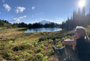a woman sitting in a prairie with a lake and mountain in the background.