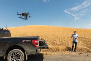 Andrew Nelson stands next to a black pickup truck parked next to a wheat field and flies a drone over the bed of the truck.