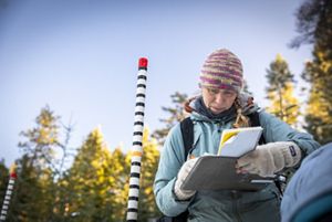 Dr. Emily Howe, TNC ecologist, wears a knit hat and winter clothing and looks at a notebook while standing in a snowy forest.