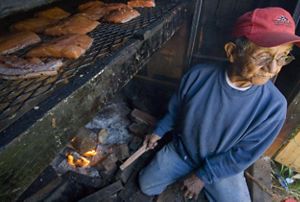 A man sits near a small fire and cooks salmon.