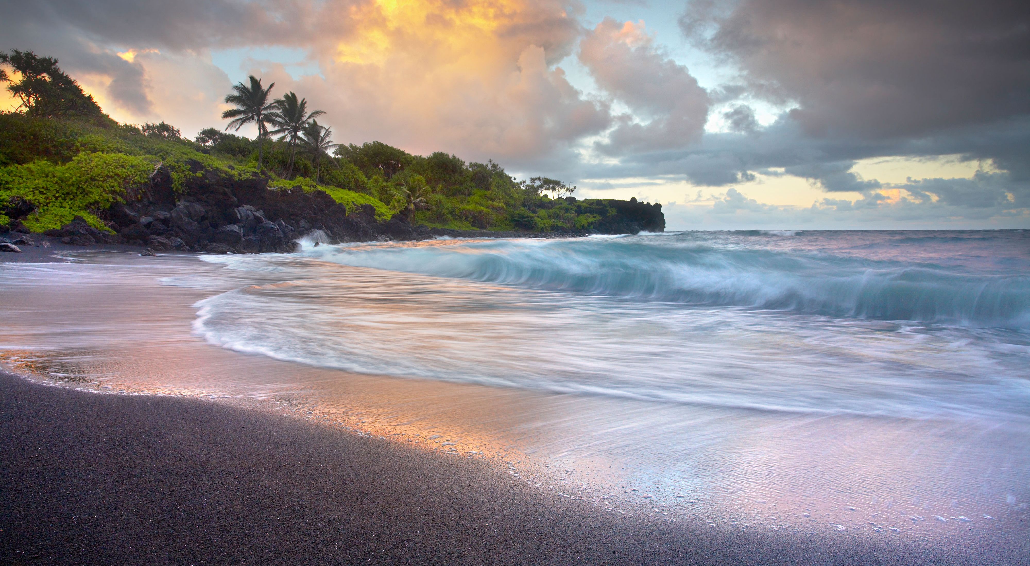 Waves crash on the black sand beach of Waianapanapa Sands with a rocky outcrop topped with lush greenery in the distance.