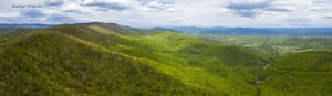 Drone view looking out over rolling mountain ridges and valleys. The mountain sides are thickly forested. Sunlight dapples over the trees. Small clearings and farms are visible in the distance.