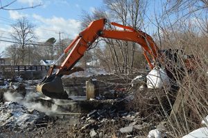 An orange excavator arm poised above the concrete remains of the West Britannia Dam on a winter day.