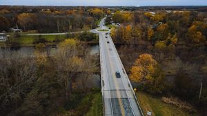 The White River in east central Indiana. Cars drive on a two-lane bridge that spans a narrow river. Buildings peek from beneath the trees that grow along the river bank.