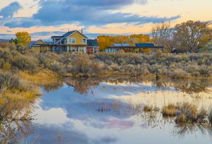 A yellow and red building reflected in a silver body of water with trees along its banks. 