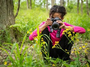 A young girl sits on the ground to take a photo of wildflowers.