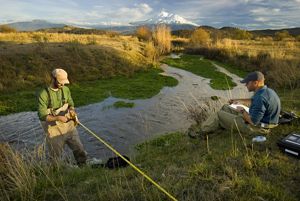 Two researchers along the Shasta River. One is standing in the river with a tape measure and the other is sitting on the bank with a clipboard.