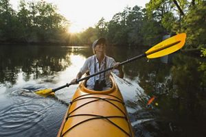 A woman paddles in a kayak along the Black Mingo Creek. There is a sunrise peeking through the treetops in the background.