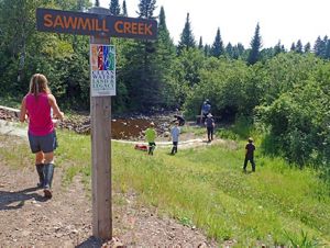 A group of people walk past a sign down a grassy hill towards a creek.