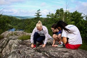 Three people crouch on a rocky outcropping overlooking a lake and forests.