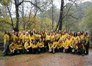 Group photo of several dozen women taking during a fire training exchange. The women are wearing yellow fire gear and posing in a clearing in a forest.