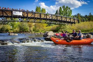 People in an orange inflatable boat on a river. A bridge with onlookers are above them.