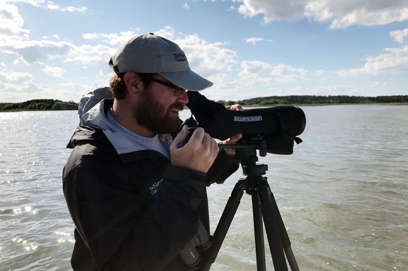 Candid snapshot of Coastal Biologist Zak Poulton. A bearded man looks through a monocular scope mounted on a tripod to observe birds at the Virginia Coast Reserve during TNC's 2017 Bird Blitz event.